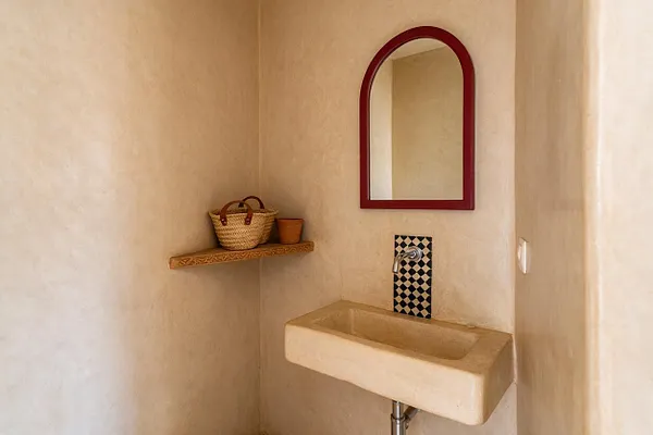 A small Moroccan-style powder room with smooth beige plaster walls, a narrow rectangular pedestal sink, and a red-framed arched mirror above it. A black-and-white patterned tile panel surrounds the faucet, and a floating wooden shelf holds a woven basket and a small terracotta cup.