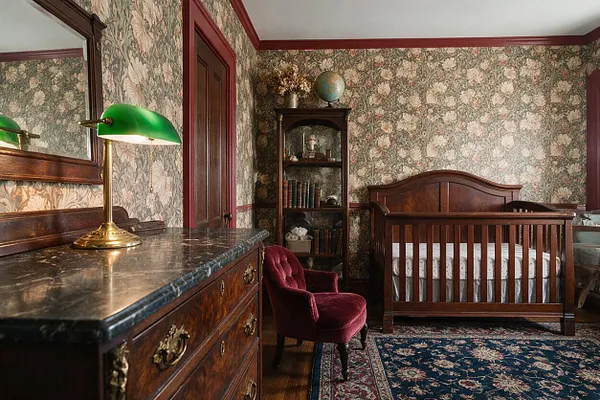 A nursery styled in dark academia features a dark wood crib, floral wallpaper, and rich burgundy trim. A vintage dresser with a green banker’s lamp, a velvet accent chair, and a patterned rug complete the moody, scholarly atmosphere.