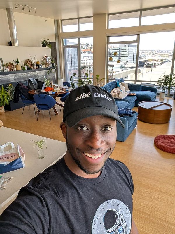 A smiling individual wearing a 'vibe coder' cap poses in a stylish, well-lit living space.
