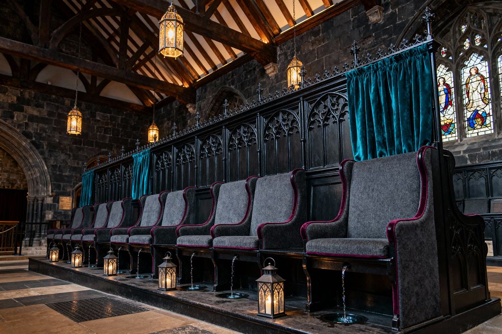 A dramatic media room styled like a chapel, with a long row of high-back gray seats trimmed in burgundy, built into black carved wood paneling with pointed-arch tracery. Teal velvet curtains, candlelit floor lanterns, and brass pendant lights illuminate dark stone walls, exposed timber beams, arched openings, and stained-glass windows over a checkerboard stone floor.