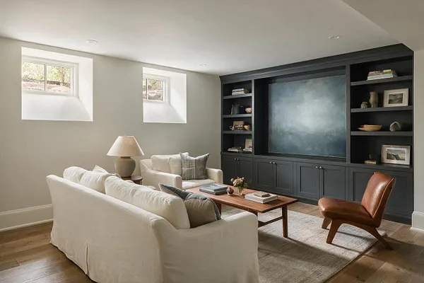 A bright transitional basement lounge with a white slipcovered sofa, cream armchairs, a wood coffee table, and a caramel leather accent chair. Dark built-in cabinetry and shelves frame a large central art panel, while two small basement windows and recessed ceiling lights keep the room feeling open and inviting.