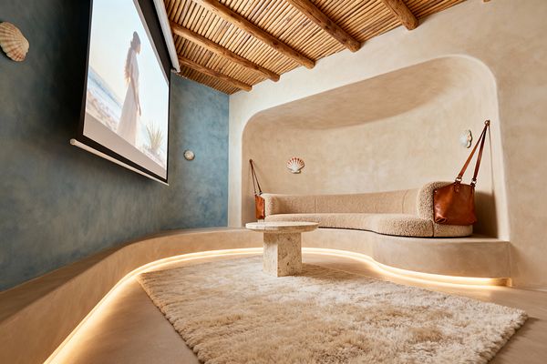A cozy media room features rounded sand-colored plaster walls forming a built-in curved sofa upholstered in beige bouclé beneath a ceiling of exposed wood beams and reed slats. A sea-blue limewash accent wall frames a drop-down projection screen, a travertine pedestal table rests on a plush cream rug, and concealed LED lighting glows under the seating plinth, with small shell accents reinforcing the coastal theme.
