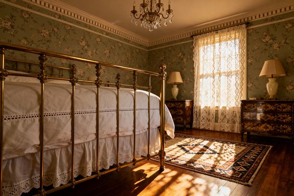 Traditional bedroom featuring a polished brass bed with white eyelet bedding, set against sage green floral wallpaper. Lace curtains filter sunlight through a single window, casting patterned shadows on warm hardwood floors and a cream, navy, and rust wool rug. Two matching inlaid wood dressers flank the window, each topped with a ceramic lamp. A crystal chandelier hangs from a ceiling detailed with dentil crown molding.