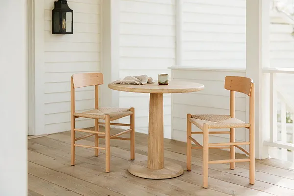 A bright Scandinavian-style porch with white horizontal shiplap walls and wide wood plank flooring. Two light oak chairs with woven seats flank a round pedestal table, creating a simple natural seating nook beside a black wall lantern.
