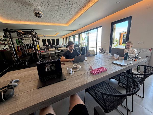 A vibrant coworking space with a large rectangular wooden table surrounded by people working on laptops under modern pendant lighting.