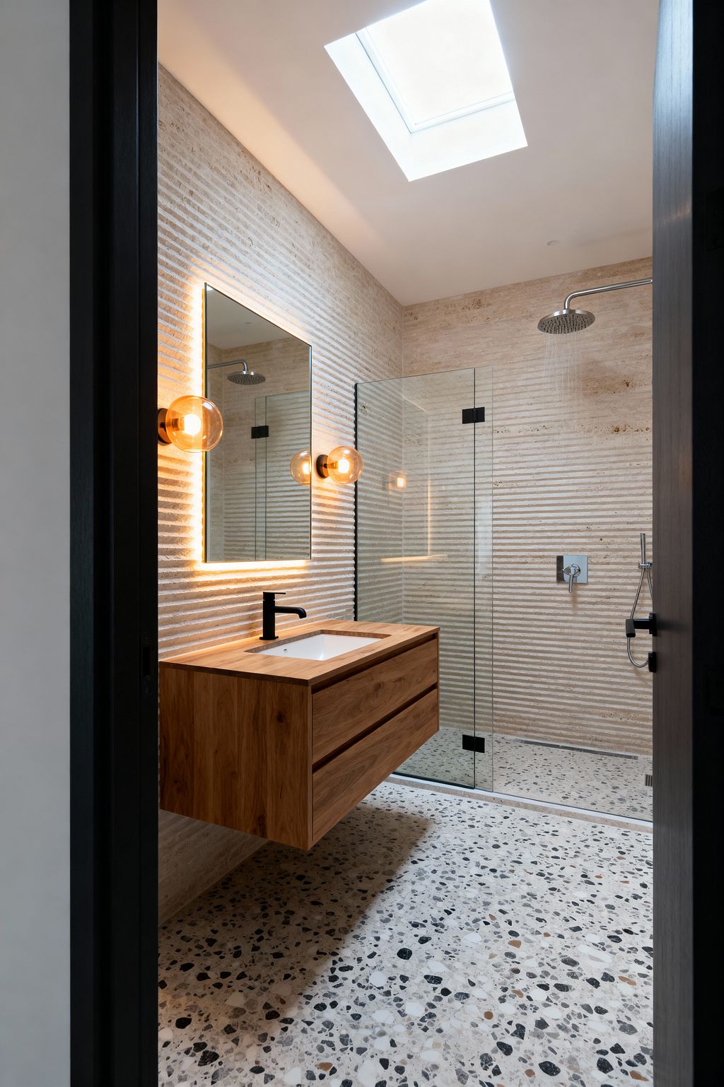 A compact contemporary bathroom with ribbed beige stone walls, a floating natural oak vanity with a white rectangular sink and matte black faucet, and a frameless glass walk-in shower with a stainless steel rain head and hand shower. A backlit rectangular mirror and two amber glass globe sconces provide warm light, while a skylight floods the terrazzo floor and curbless shower with daylight for a seamless, open feel.