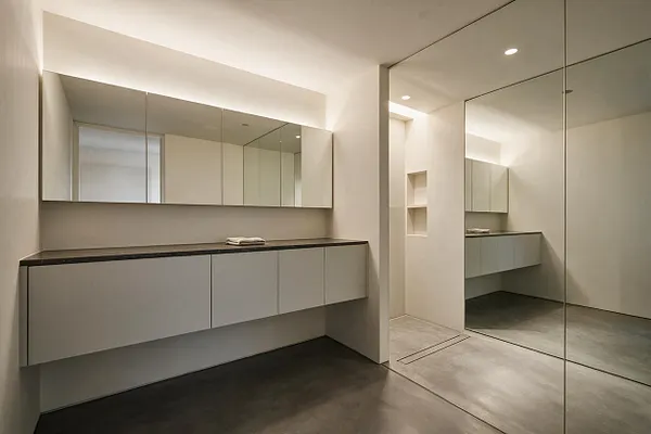 A minimalist master bathroom with a floating vanity, long mirrored medicine cabinets, and a dark stone countertop. Large floor-to-ceiling mirrored panels reflect the room, while soft recessed lighting and pale walls create a calm, spacious feel.