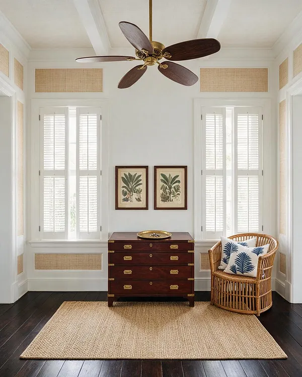 A bright tropical-style entryway with white walls, tall shuttered windows, and woven wall panels. A dark wood chest with brass hardware sits beneath framed botanical prints, beside a rattan chair with patterned pillows and a large natural fiber rug.