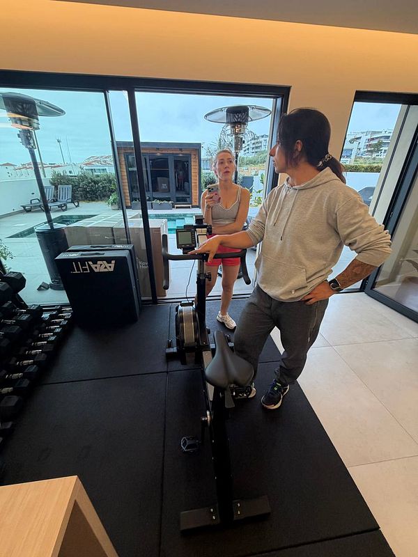 A woman in gym attire relaxing on a black couch while holding a phone and a drink in a gym setting.