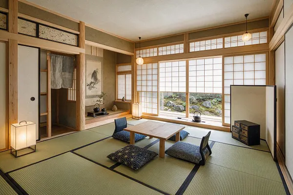 A Japanese Zen basement room with tatami mat flooring, low wooden table, floor cushions, and shoji screen windows along the back wall. Soft pendant lights, sliding doors, and a small alcove with minimalist decor create a calm, traditional atmosphere.