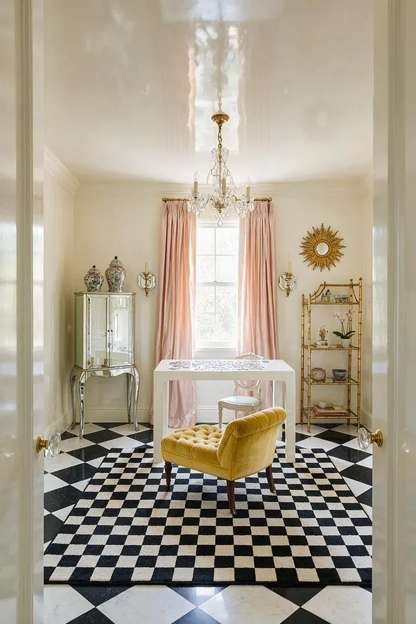 A refined game room features a glossy white ceiling, a crystal chandelier, blush pink drapes framing a bright window, and a black-and-white checkered rug over a marble-like diamond floor. A mustard velvet chair sits in front of a sleek white game table, with mirrored and brass accent furniture, a sunburst mirror, and decorative ceramics completing the glamorous look.