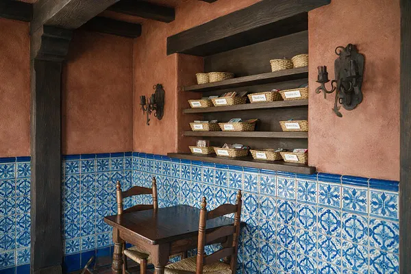 A small Mediterranean-style game room features terracotta stucco walls, dark exposed ceiling beams, and a band of blue-and-white patterned tile wrapping the lower walls. A dark wood table with two woven-seat chairs sits in the corner, while open shelves hold wicker baskets and small labeled items beneath wrought iron wall sconces.