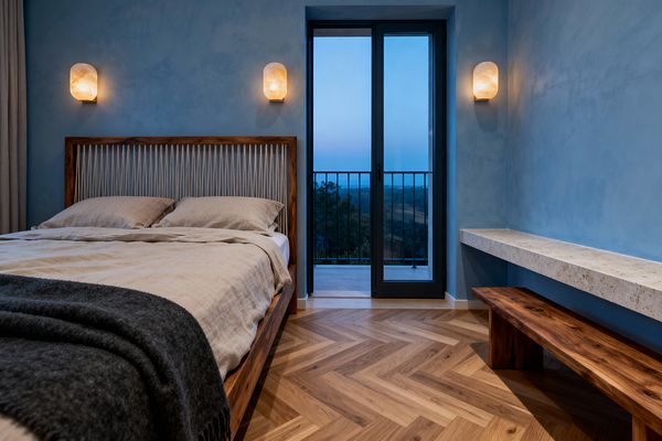 Scandinavian bedroom with dusty blue limewash walls and warm oak herringbone flooring features a walnut platform bed with a rope-laced headboard, beige linen bedding, and a charcoal wool throw. Two glowing alabaster sconces flank a black metal-framed glass door to a small balcony, while a floating travertine ledge and a simple walnut bench line the right wall, with neutral drapery softening the left side for a calm, minimal atmosphere.
