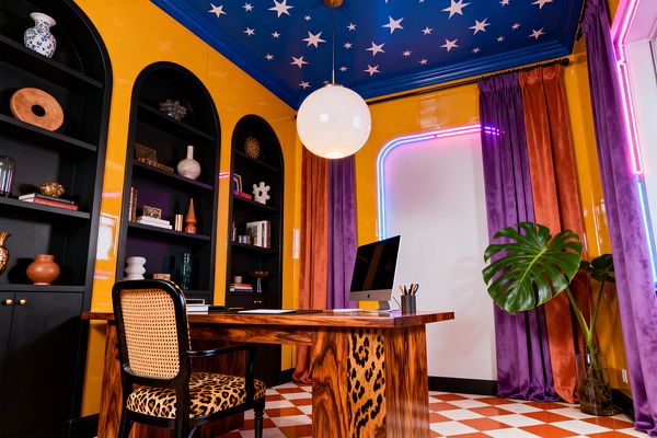 A bold home office features marigold walls and black arched built-in shelves filled with pottery and books. A cobalt blue ceiling painted with white stars hangs a globe pendant above a highly figured burl wood waterfall desk and a black chair with a cane back and leopard-print seat; purple and rust velvet curtains frame a corner traced by pink-to-blue neon, while a terracotta-and-white checkerboard floor and large monstera leaves complete the vivid maximalist scene.