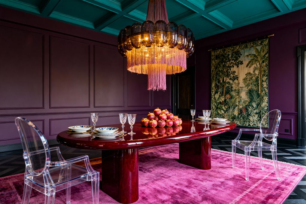 A dramatic dining room with deep plum paneled walls and a teal coffered ceiling features a large oval burgundy lacquer table set with crystal stemware and blue-and-white china. Clear acrylic armchairs sit on a saturated pink rug over a black chevron wood floor. Overhead, a chandelier of smoked glass and warm bulbs is finished with mauve fringe. A verdant landscape tapestry hangs on the right wall; a mound of pomegranates forms the centerpiece.