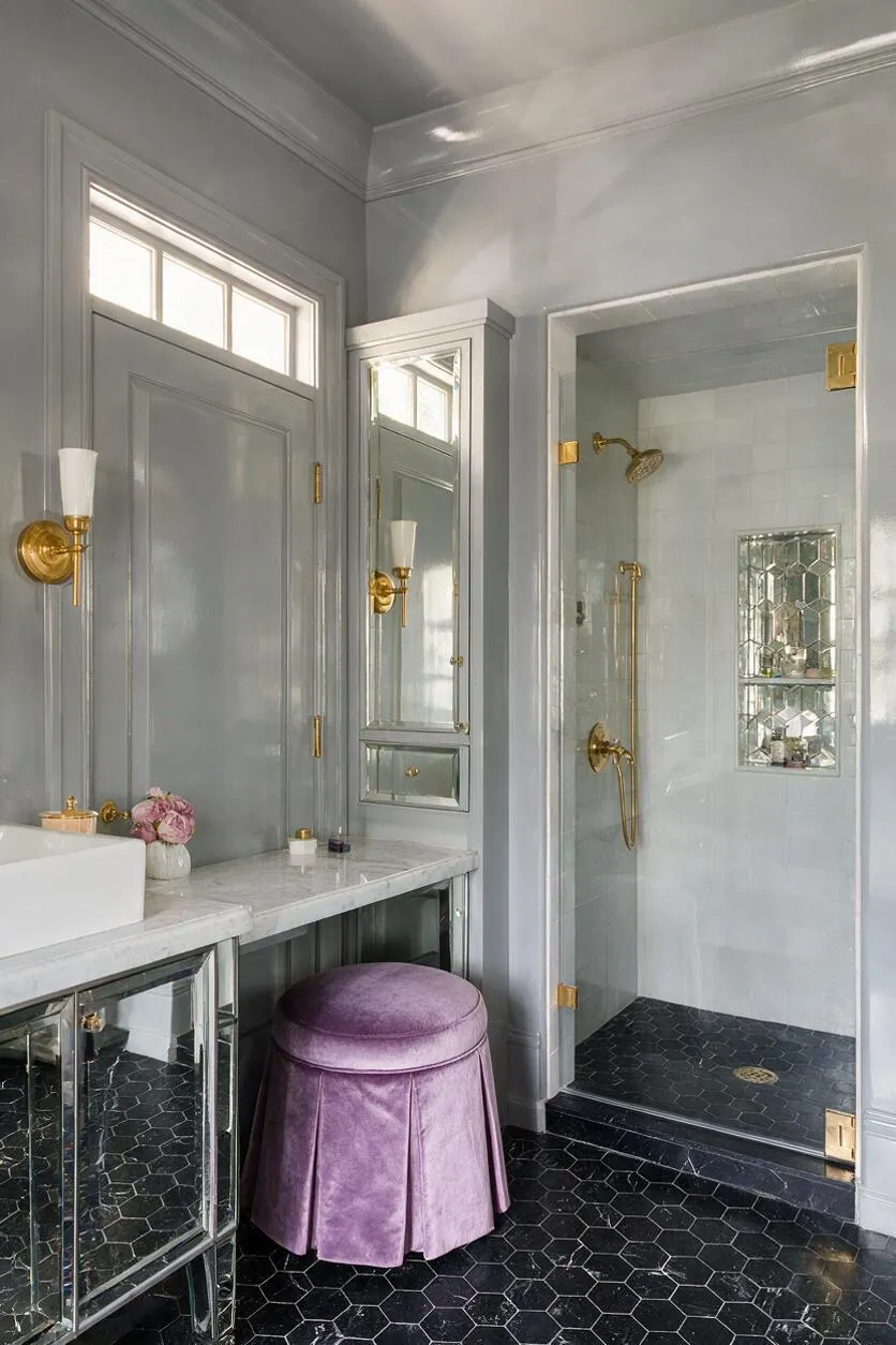 A Hollywood Regency master bathroom with glossy gray walls, a marble vanity, mirrored cabinet doors, and brass sconces and shower fixtures. The shower has white tile walls, a small mirrored niche, and a dark hexagonal tile floor, while a lavender velvet stool adds a soft decorative accent.