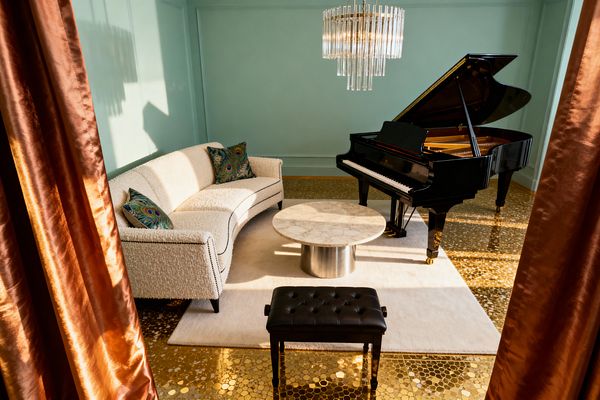 A mint-walled living room centers on a glossy black grand piano beside a curved ivory bouclé sofa with contrast piping and peacock-pattern pillows, arranged around a round marble coffee table on a brushed metal base and a plush ivory rug. Warm light filters through copper satin drapes onto a sparkling gold mosaic floor, while a tiered crystal-rod chandelier hangs above and a tufted black bench stands in the foreground.