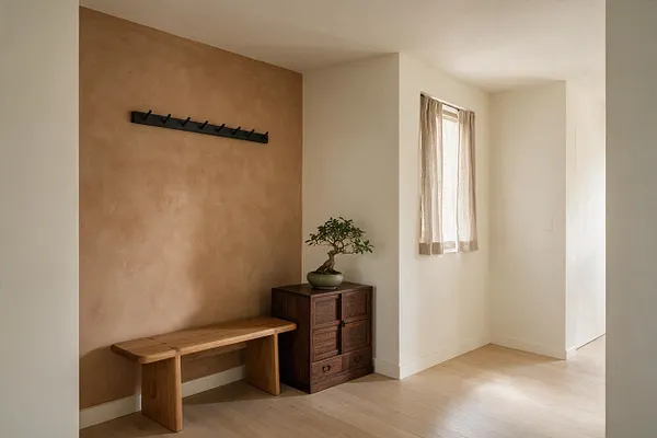 A serene Japandi entryway with a warm beige textured accent wall, a black wall-mounted coat rack, and a simple wooden bench beneath it. A small dark wood cabinet topped with a bonsai-style plant sits beside a softly lit window with sheer curtains, and pale wood flooring keeps the space calm and minimal.