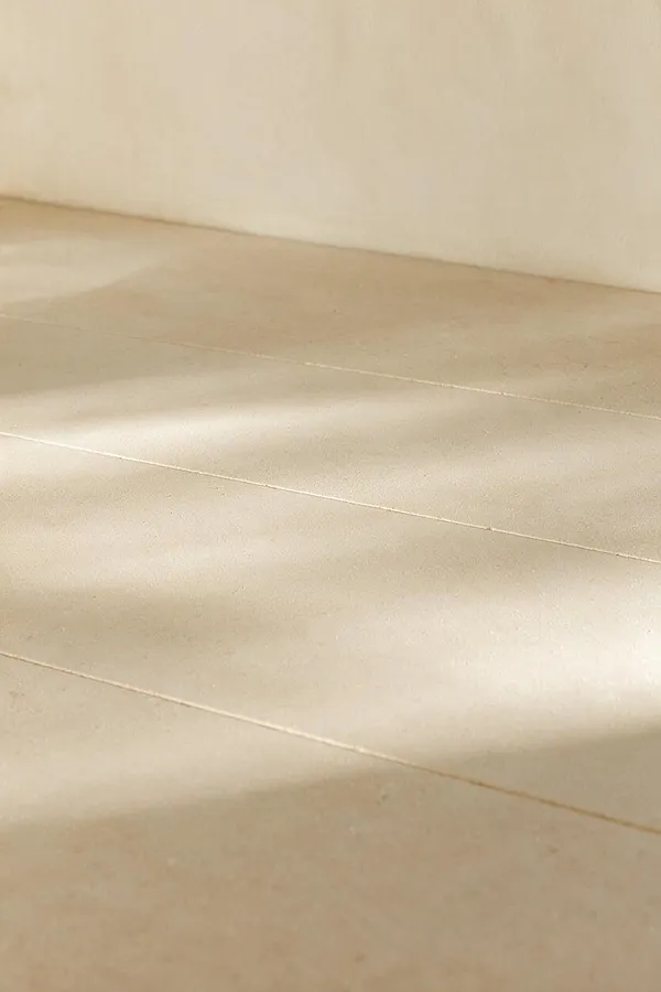 A close-up view of a minimalist interior floor made of large beige stone or concrete tiles with thin grout lines. Soft natural light creates gentle highlights and shadows across the smooth surface, and a plain light wall meets the floor at the top of the image.