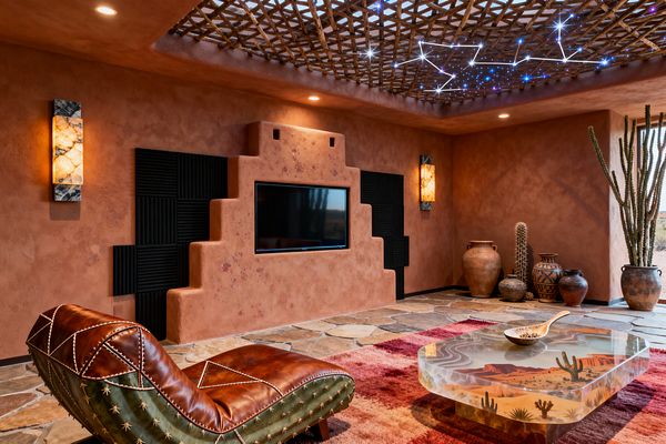 A Southwestern media room with adobe-textured terracotta walls features a stepped TV niche flanked by black acoustic panels and glowing onyx sconces. A twig-lattice ceiling with fiber‑optic constellations hangs over flagstone floors, a striped red rug, a resin coffee table depicting a desert landscape, and a saddle-brown leather chaise; large earthenware pots and tall cacti anchor the corner beside a window to the desert.