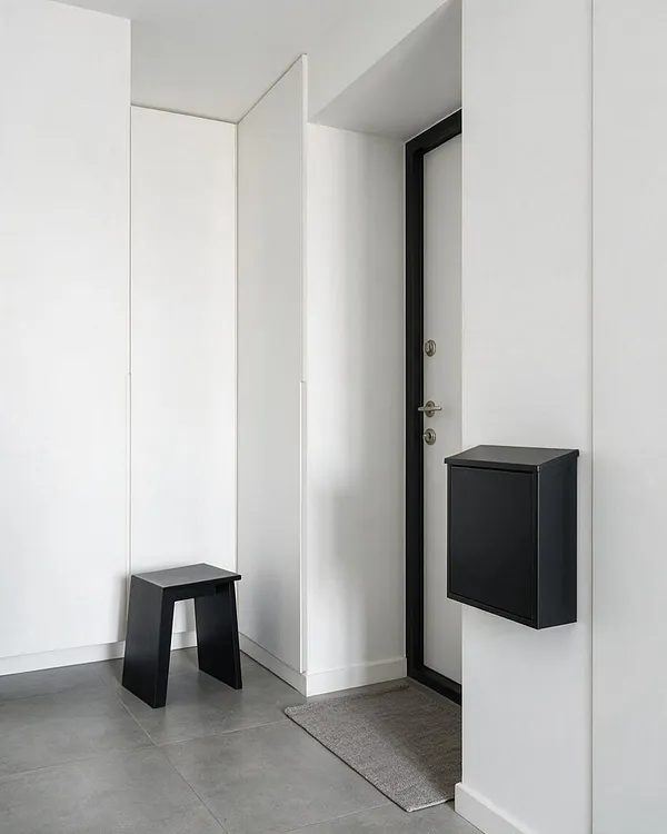 A minimalist front porch with smooth white walls, large gray floor tiles, and a black-framed front door. A small black stool sits in the corner, and a sleek black wall-mounted mailbox and gray doormat add simple, functional details.