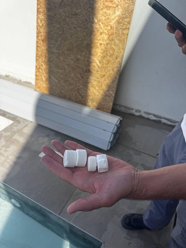 A hand holds three white chemical tablets against a backdrop of a pool area.