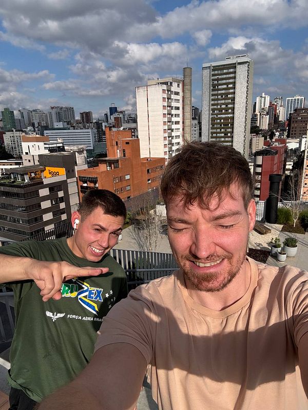 Two men take a selfie on a rooftop with a city skyline in the background.