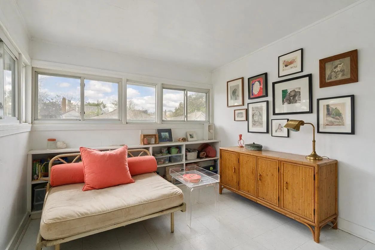 A bright sunroom with white walls, a row of windows, and a simple daybed upholstered in beige with coral pillows. Along one wall are open shelves and a honey-toned wood sideboard topped with a brass lamp, with an eclectic gallery wall of framed art above it and a clear acrylic table in the center.
