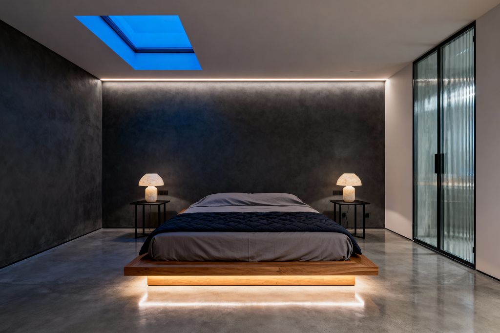 A modern bedroom features a low oak platform bed with underbed LED lighting that creates a floating effect. Charcoal plaster walls with continuous cove lighting, polished concrete floors, and two black side tables topped with glowing alabaster mushroom lamps frame the scene, while a blue-tinted skylight and fluted glass sliding doors on the right add cool contrast and soft privacy.