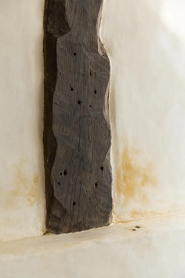 A close-up of a dark, weathered wooden beam set into a cream plaster wall. The rough wood grain, small knot holes, and aged patina contrast with the soft, warm-toned plaster surrounding it.