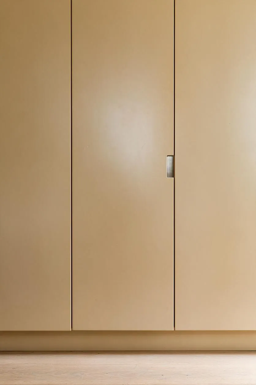 A close-up view of flat-panel beige cabinet doors in a minimalist interior, with slim vertical seams and a small recessed metal handle on one door. The cabinets sit above a light wood floor, creating a calm neutral palette.