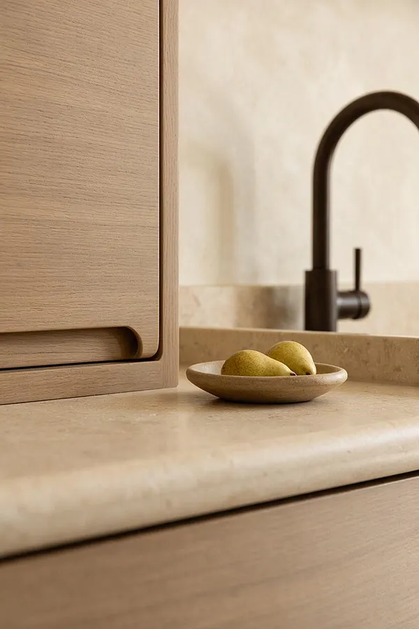 A close-up of an Organic Modern vanity detail with light oak cabinetry, a creamy stone countertop and backsplash, and a matte black arched faucet. A small shallow bowl holding two pears sits on the counter, adding a natural touch.