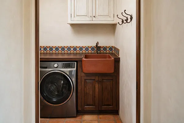 A narrow Mediterranean-style laundry room with a front-loading washer built into dark wood cabinetry beside a deep terracotta sink. The space features a colorful patterned tile border, terracotta floor tiles, white plaster walls, and a small white wall cabinet above.