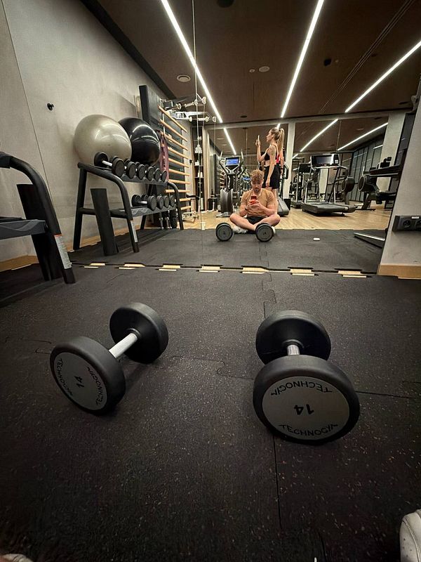 A gym room with workout equipment, two dumbbells in the foreground, and a mirrored wall reflecting a person sitting on the floor and another standing.