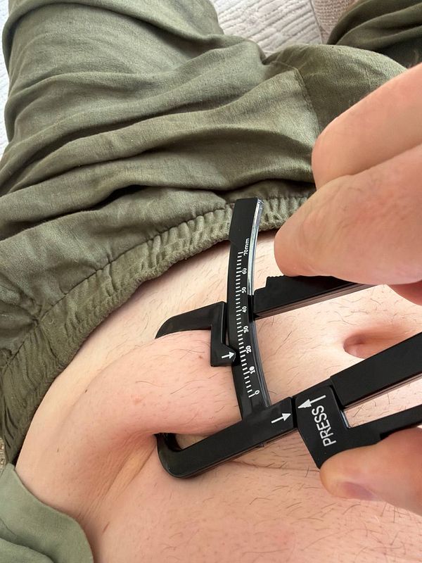 Hand holding a skinfold caliper measuring abdominal fat on a person's belly, with a digital kitchen scale and black tape measure in the background.