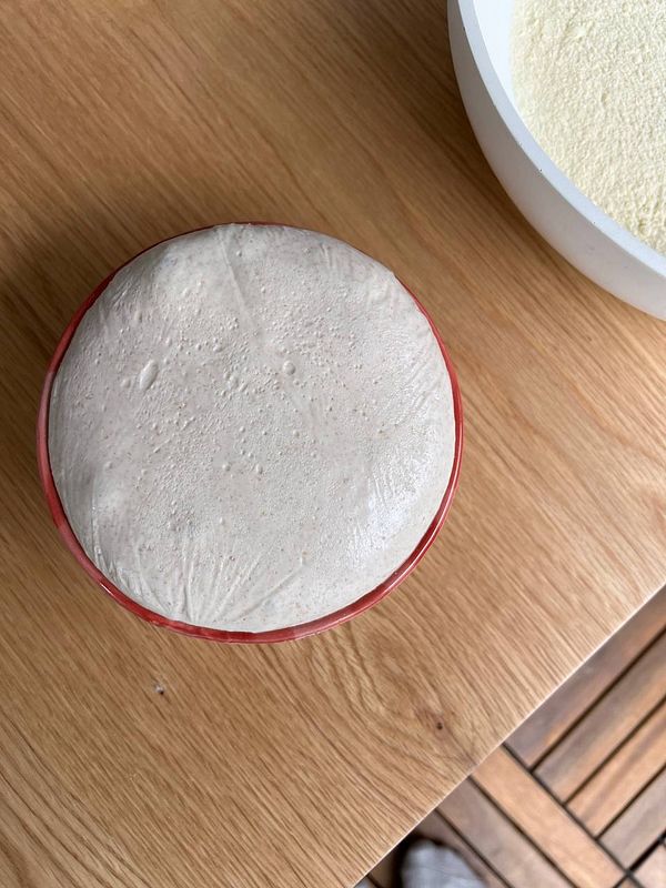 A bowl of risen dough sits on a wooden surface next to a larger bowl of flour.