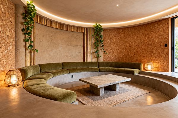 A circular, sunken media room features a built‑in curved sectional upholstered in olive‑green velvet surrounding a low rectangular stone coffee table on a jute rug. Walls combine textured clay plaster, bamboo slat accents, and warm OSB panels, with trailing pothos plants adding greenery. A continuous cove light glows around the ceiling, two rattan lanterns sit at the edges, and a sliding glass door opens to the outdoors.