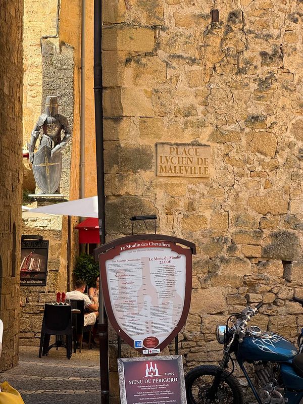 A picturesque medieval village scene featuring a knight statue, a restaurant menu, and a vintage motorcycle.