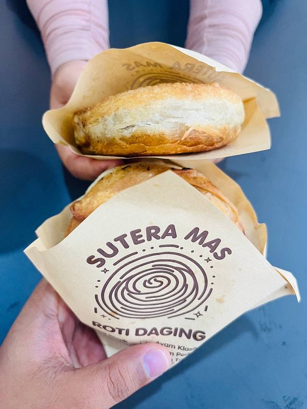 Two pieces of roti daging are being held in paper wrappers.