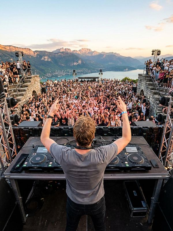 A DJ performs in front of a large crowd at an outdoor music festival with a scenic backdrop.