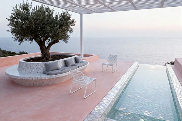 A minimalist coastal patio with a blush-pink concrete floor faces an open sea view. A curved terrazzo planter-bench with an olive tree and gray cushions sits beside a slim lap pool lined in iridescent glass mosaic, while two white slatted metal chairs rest under a white louvered pergola casting soft linear shadows; the overall scene is calm, sunwashed, and contemporary.