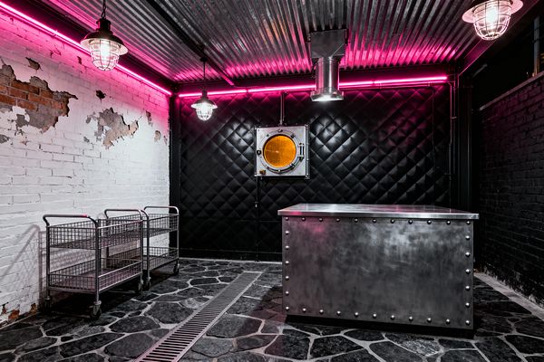 An industrial laundry room with a corrugated metal ceiling and cage-shaded pendants under a perimeter of magenta neon. A glossy black, diamond-quilted wall with a round amber porthole contrasts distressed white brick; a riveted stainless-steel island sits on dark flagstone with a linear floor drain, while two chrome wire rolling carts line the left wall.