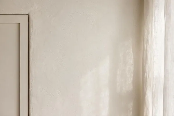 A close-up interior view of a softly textured beige plaster wall beside a simple framed panel on the left and sheer off-white curtains on the right. Natural daylight filters across the surface, creating a warm, calm Transitional look.