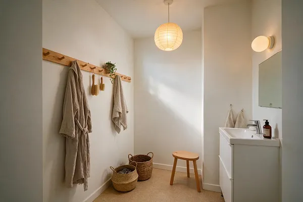 A bright master bathroom with white walls, a small vanity on the right, and a round paper pendant light centered overhead. Natural wood accents include a wall-mounted coat rack, a small stool, and woven baskets, while beige towels add a soft, calming texture.