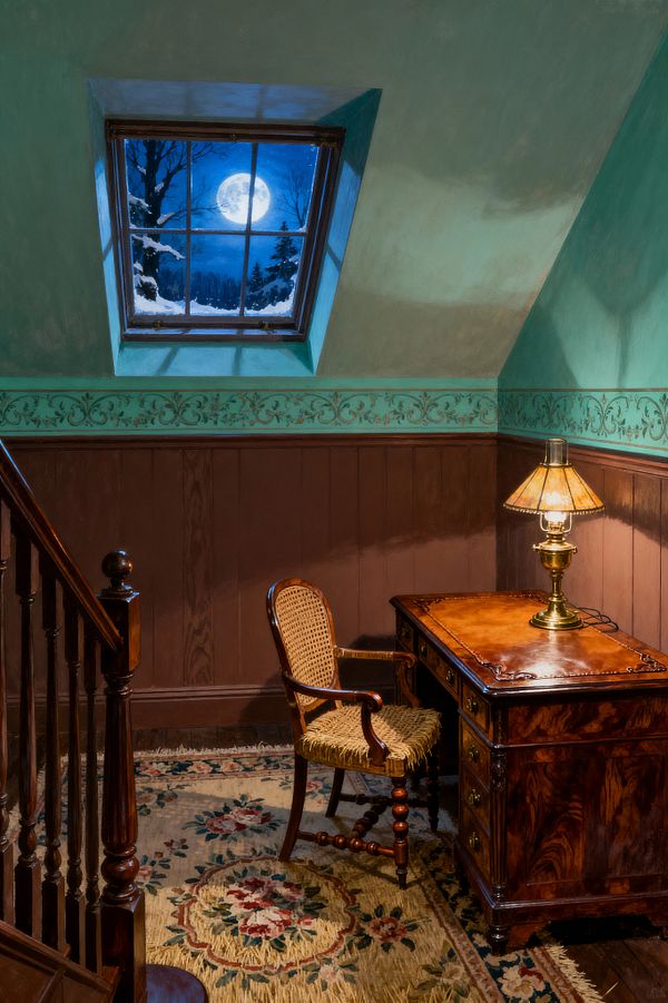 A compact Victorian-style home office sits beneath a sloped ceiling, where a skylight frames a snowy night and full moon. Dark wood wainscoting and staircase contrast mint green walls with a stenciled frieze; an antique mahogany desk with a tooled leather top and brass pulls holds an amber-glass table lamp, while a wooden chair with caned back and rush seat rests on a cream floral wool rug.