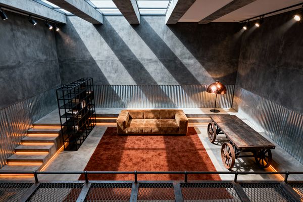A sunken industrial living room features raw concrete walls, corrugated metal cladding, and a skylit ceiling with concrete beams casting dramatic diagonal shadows. At the center sits a camel suede sofa on a rust-colored rug, flanked by a black metal open bookshelf and a vintage-style table made from weathered wood on iron wheels, with linear amber LED lighting tracing the steps and perimeter and a dome floor lamp adding a metallic glow.