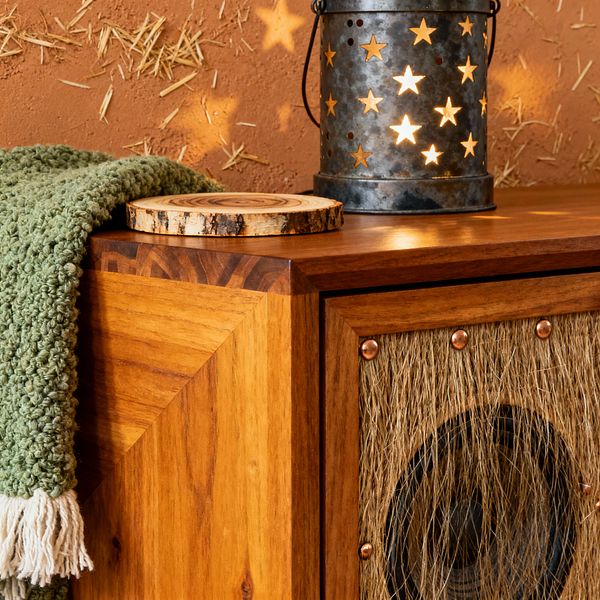 Close-up of a rustic media console made of honey-toned wood with mitered corners. A speaker sits behind a jute fiber grille secured with copper rivets. On top, a chunky sage green knit throw with cream fringe lies beside a live-edge wood slice. A galvanized metal lantern with star cutouts casts warm light and star-shaped shadows across a terracotta, straw-flecked plaster wall, creating a cozy, handcrafted atmosphere.