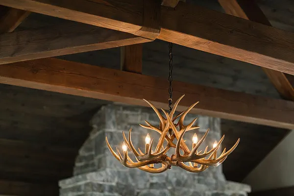 A rustic interior ceiling with heavy exposed wooden beams and a hanging antler-style chandelier centered in the frame. The fixture has multiple candle-like bulbs and sits in front of a stone fireplace or chimney surround.