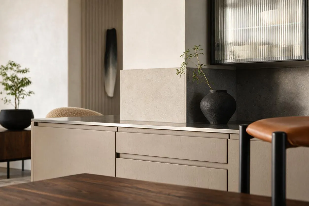A modern kitchen detail with beige flat-panel cabinets, a dark countertop, and a light stone backsplash beneath ribbed glass upper cabinets. Black ceramic vases, warm wood furniture, and a tan leather stool add contrast to the calm neutral palette.