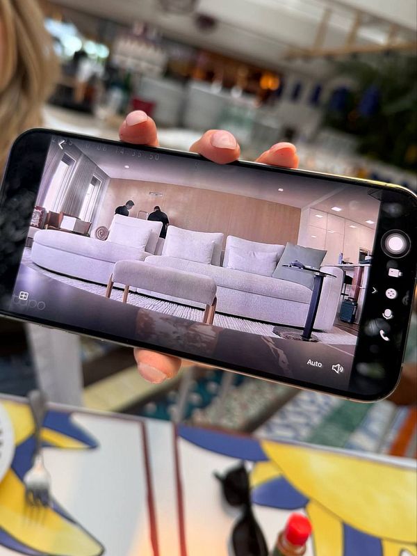 A smartphone displays a remote view of a living room with a large white sofa and two people present.
